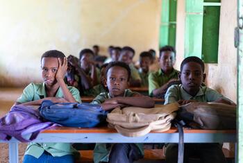 A group of young school children in green uniforms sit at a wooden desk in a classroom.