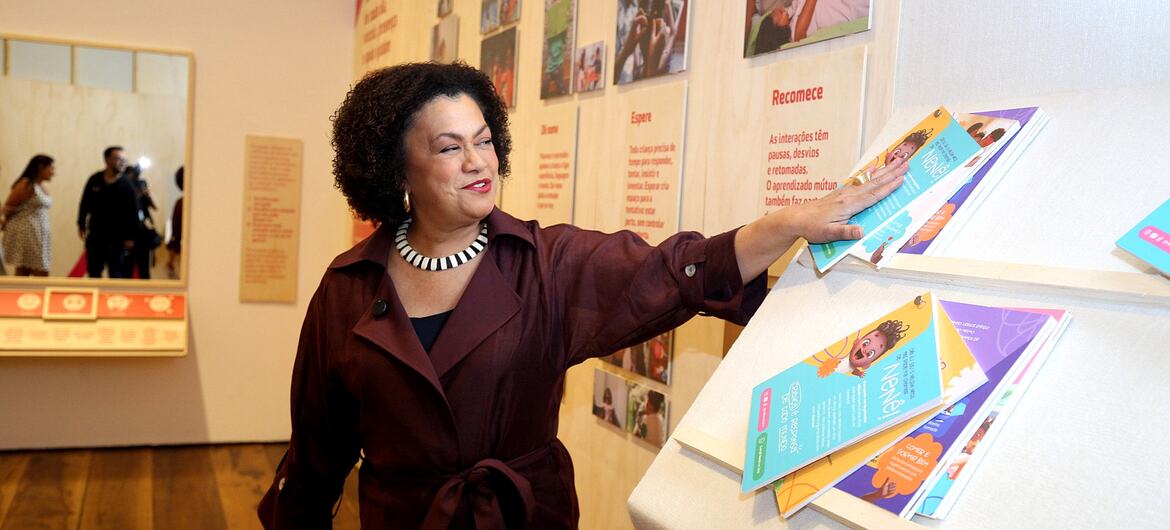 Maria Angela de Jesus está em frente a uma vitrine, apontando para uma pilha de livros infantis coloridos.