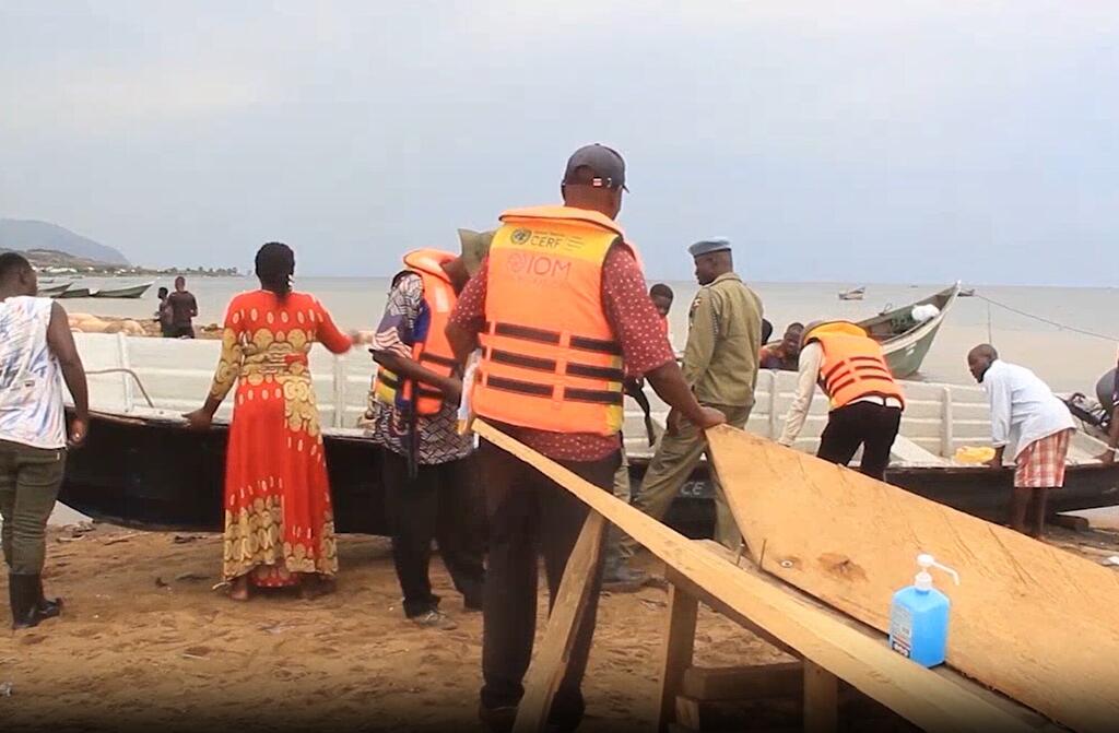 Kundi la watu waliovaa fulana za kuokoa maisha za rangi ya chungwa wakifanya kazi kwenye mashua ya mbao kwenye ufuo wa mchanga nchini Uganda. Mwanamke aliyevaa gauni jekundu amesimama karibu huku wanaume wakishughulikia mbao na vifaa karibu na Ziwa Albert.