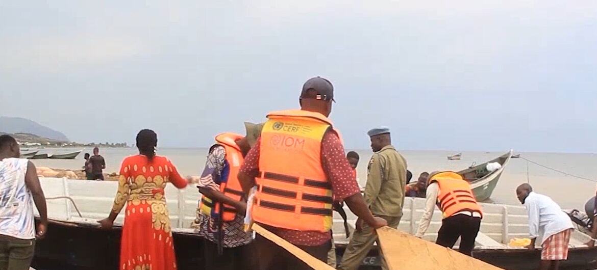 A group of people wearing orange life vests work on a wooden boat at a sandy shoreline in Uganda. A woman in a red patterned dress stands nearby as men handle planks and equipment near Lake Albert.