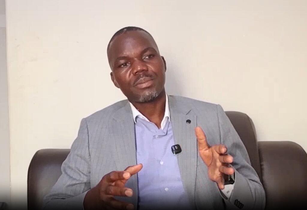 Mwanamume aliyevaa suti ya kijivu na shati la bluu ameketi kwenye sofa ya ngozi ya kahawia, akiashiria kwa mikono yake huku akiwa amevaa kipaza sauti cha begi.