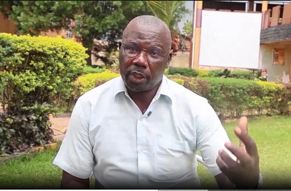 Mwanamume Mwafrika aliyevaa shati jepesi na miwani ya mviringo anazungumza nje karibu na Ziwa Albert, Uganda, akiwa na ua wa kijani uliopambwa vizuri na jengo nyuma.