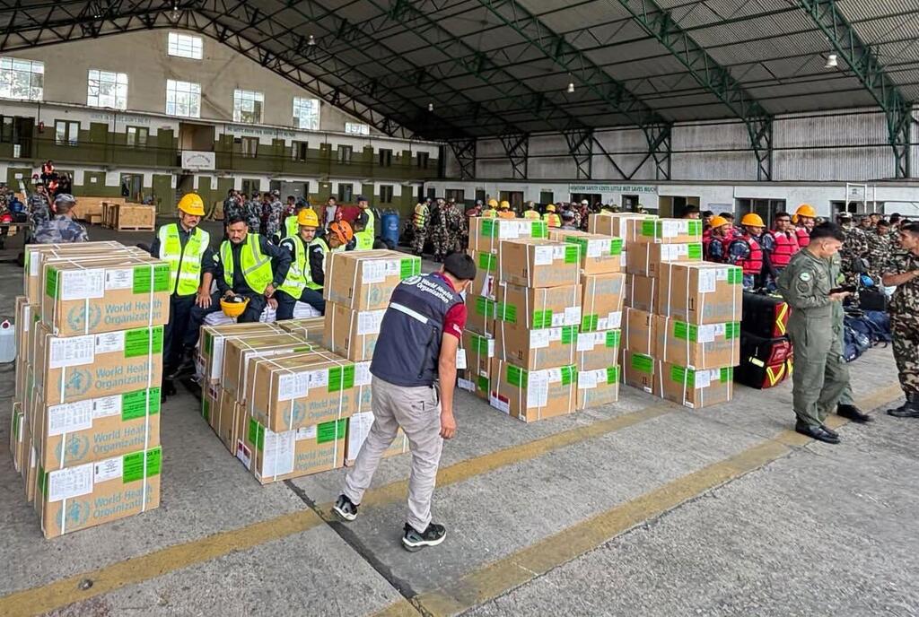 Des ouvriers portant des gilets et des casques de sécurité manipulent des caisses de ravitaillement empilées et étiquetées par l'OMS à l'intérieur d'un grand entrepôt, sous le regard de soldats postés à proximité.