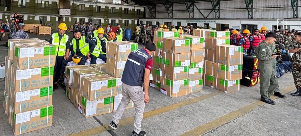 Workers in safety vests and helmets handle stacked WHO-labeled supply boxes inside a large warehouse, with soldiers observing nearby.