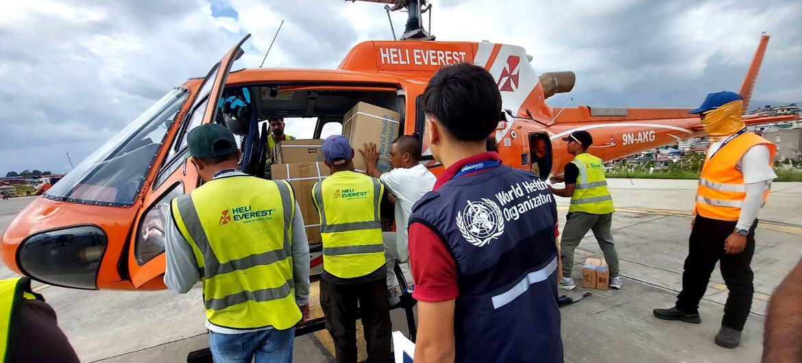 Funcionários da OMS e da Heli Everest carregam caixas de suprimentos médicos de emergência em um helicóptero laranja na pista de um aeroporto no Nepal.