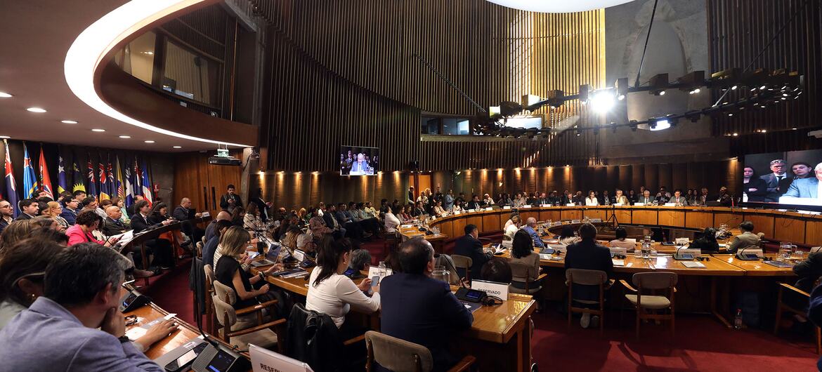 Los delegados, sentados en mesas curvas de madera en una gran sala de conferencias al estilo de la ONU durante una reunión de la CEPAL sobre inteligencia artificial, con banderas nacionales y pantallas de video que muestran a los participantes remotos.