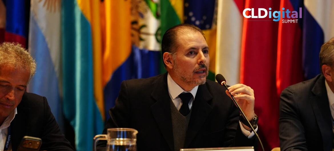 Fernando Rojas, de la Comisión Económica para América Latina y el Caribe, habla por un micrófono en un panel de la Cumbre CLDigital, sentado detrás de una placa con las banderas nacionales de los países latinoamericanos dispuestas al fondo.