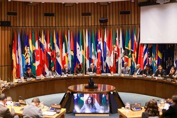 Diplomáticos sentados en una mesa curva en una sala de conferencias de las Naciones Unidas , con las banderas nacionales de los estados miembros dispuestas detrás de ellos y una pantalla que muestra a un orador remoto.