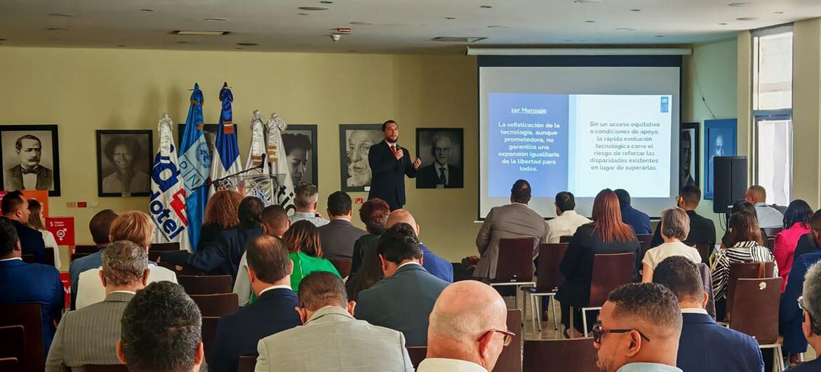Un hombre con traje oscuro se dirige a un público sentado en un evento del UNDP sobre inteligencia artificial que se celebra en un espacio cerrado, con banderas y una pantalla de presentación en español detrás de él.