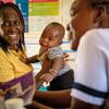 Mama Mwafrika mwenye tabasamu akiwa amevaa shati la njano amembeba mtoto wake huku akizungumza na mfanyakazi wa afya katika kliniki moja barani Afrika.