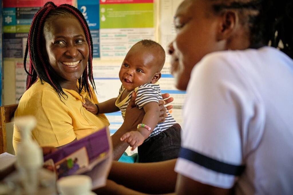 Mama Mwafrika mwenye tabasamu akiwa amevaa shati la njano amembeba mtoto wake huku akizungumza na mfanyakazi wa afya katika kliniki moja barani Afrika.