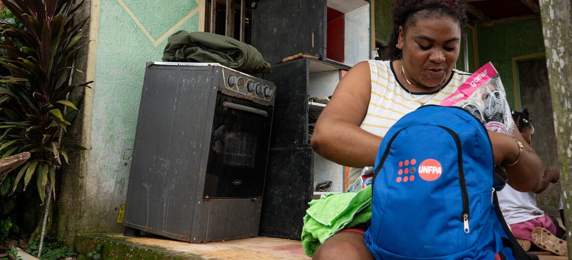 Una mujer afrocolombiana está sentada en el umbral dañado de una puerta en Chocó, organizando una mochila azul del UNFPA con artículos de higiene y dignidad.