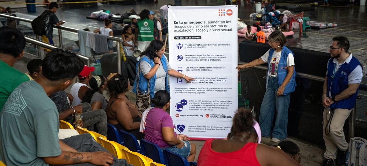 Un miembro del equipo del UNFPA , con un chaleco azul, presenta una pancarta informativa sobre los riesgos para la salud sexual y reproductiva durante la emergencia a las personas desplazadas sentadas en sillas amarillas de estadio, mientras otro miembro del personal permanece de pie cerca.