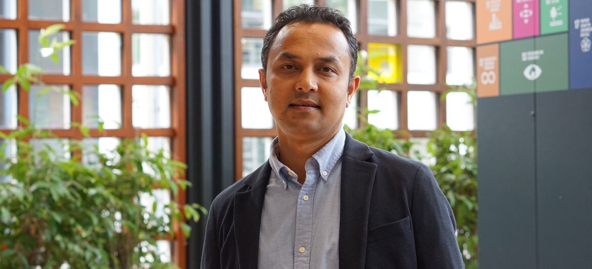 Dipesh Chapagain stands in a modern building interior with wooden lattice windows and green plants behind him.