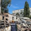 Сотрудник ВОЗ стоит среди обломков после ракетного удара, повредившего территорию детской больницы в Киеве; на фоне чистого голубого неба видны сгоревшие автомобили и обрушившиеся здания.
