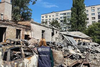 A WHO staff member stands amid the wreckage of a missile strike that damaged the grounds of a children's hospital in Kyiv, with burned-out cars and collapsed structures visible under a clear blue sky.