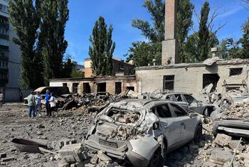 Destroyed cars and rubble in the grounds of a hospital in Kyiv after a missile attack, with several people standing nearby examining the damage.
