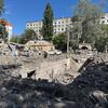 Жители осматривают большую воронку от ракетного удара и обломки на территории детской больницы в Киеве, на заднем плане — поврежденные автомобили и многоквартирные дома под ясным голубым небом.