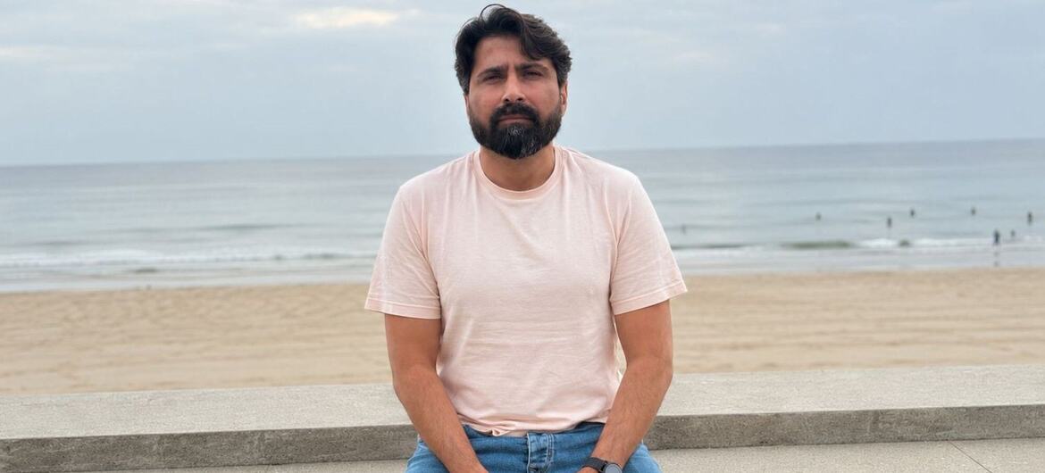 Um homem barbudo, vestindo uma camiseta rosa claro e calça jeans azul, está sentado em um degrau de granito na praia, com o mar e a areia ao fundo, sob um céu nublado.