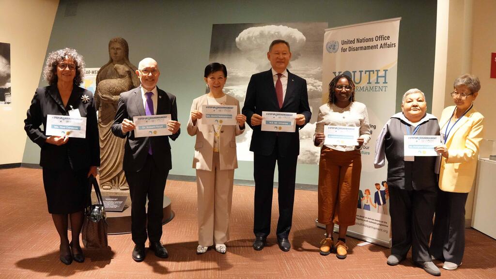 Six personnes, alignées, tiennent des certificats lors d'une exposition. Derrière elles, une grande photo en noir et blanc d'une explosion nucléaire et une banderole portant l'inscription « Bureau des affaires de désarmement des Nations Unies » avec le mot « JEUNESSE » visible.
