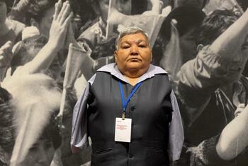 An elderly Japanese woman with short grey hair wearing a dark vest over a light shirt and a blue lanyard with an ID badge stands in front of a large black-and-white mural depicting crowds of people with raised arms.