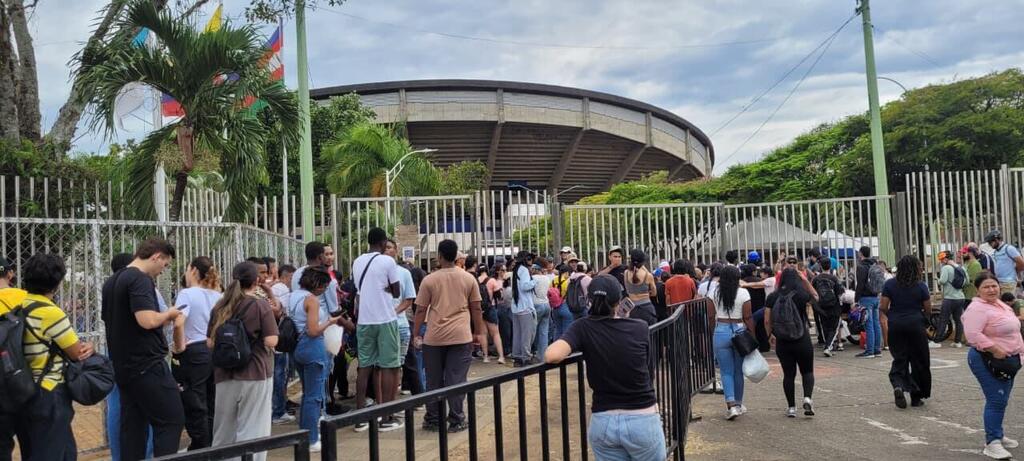 Une foule nombreuse fait la queue devant une grille métallique, face à un stade moderne.