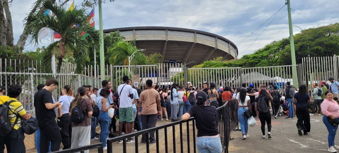 A large crowd of people stands in a long queue outside a metal gate in front of a modern stadium.