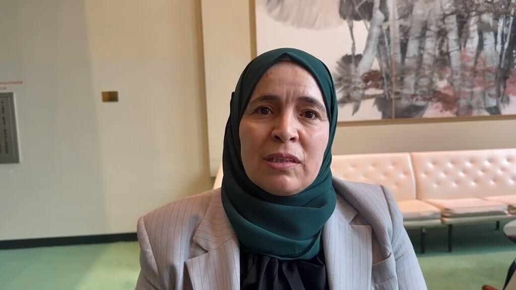 امرأة ترتدي حجابًا أخضر داكنًا وسترة رمادية تقف في الداخل، وخلفها مقعد كريمي اللون منجد ولوحة جدارية تجريدية.