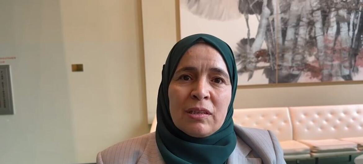 A woman wearing a dark green hijab and grey blazer stands indoors, with a tufted cream bench and abstract wall art behind her.
