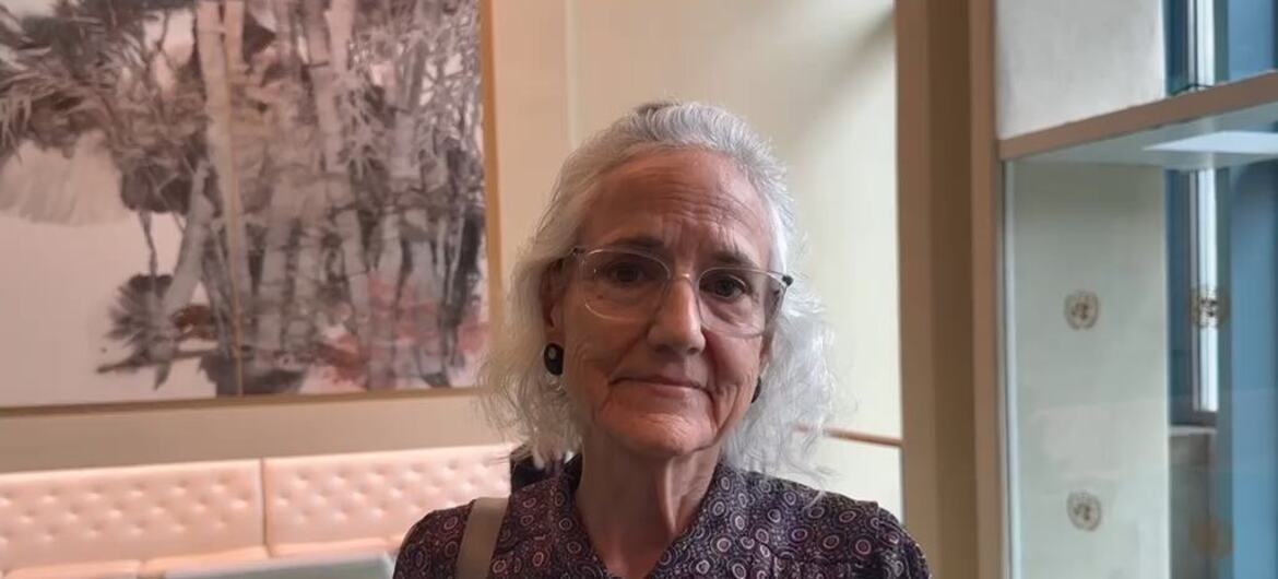 An older woman with white hair and glasses wears a patterned dark blouse, standing indoors beside a tufted bench and a large framed artwork.