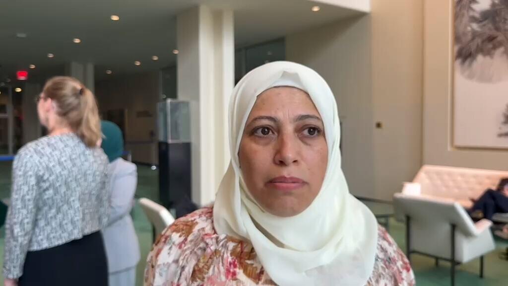 امرأة ترتدي حجاباً أبيض وقميصاً مزهراً تقف في منطقة جلوس، مع وجود أشخاص جالسين وكراسي مبطنة في الخلفية.