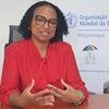 Uma mulher de blusa vermelha e óculos está sentada em uma mesa, com uma placa da Organização Mundial da Saúde referente a Moçambique na parede atrás dela.