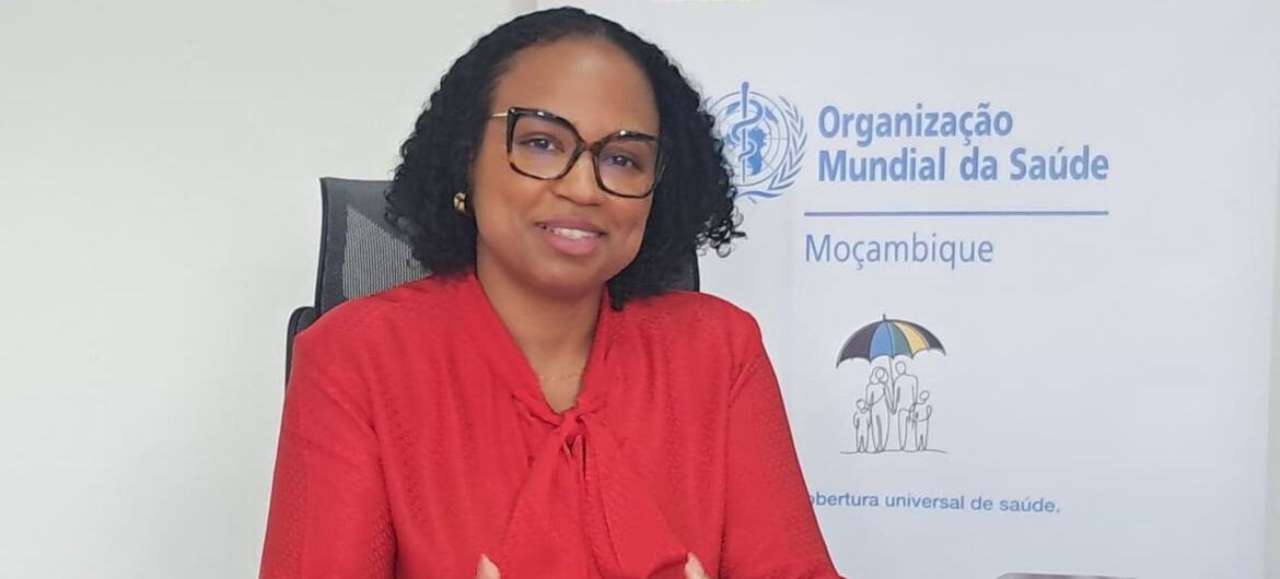 Uma mulher de blusa vermelha e óculos está sentada em uma mesa, com uma placa da Organização Mundial da Saúde referente a Moçambique na parede atrás dela.