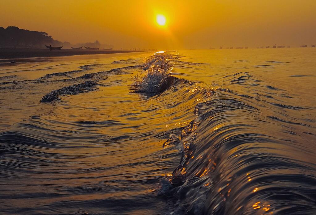 Vue en contre-plongée des vagues de l'océan reflétant le soleil couchant sur un ciel doré près d'une côte du Bangladesh, avec de petits bateaux visibles au loin sur le rivage.