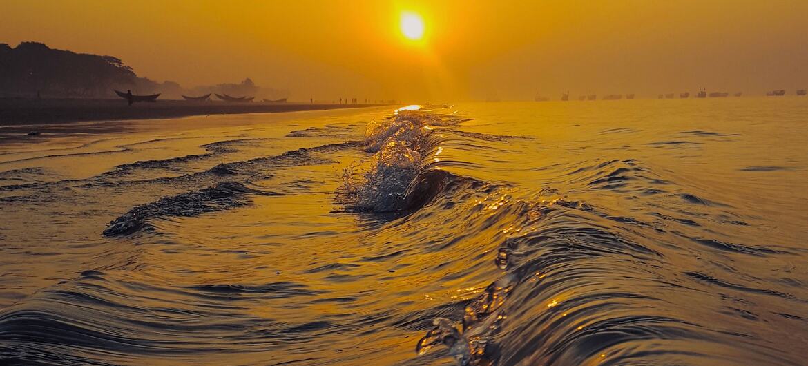 Low-angle view of ocean waves reflecting the setting sun on a golden sky near a coastline in Bangladesh, with small boats visible along the distant shore.