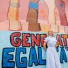 Une jeune femme blonde vêtue d'une robe bleu clair se tient devant une fresque murale colorée de street art portant l'inscription roumaine « GENERATEIA EGALAȚII » (Génération Égalité).