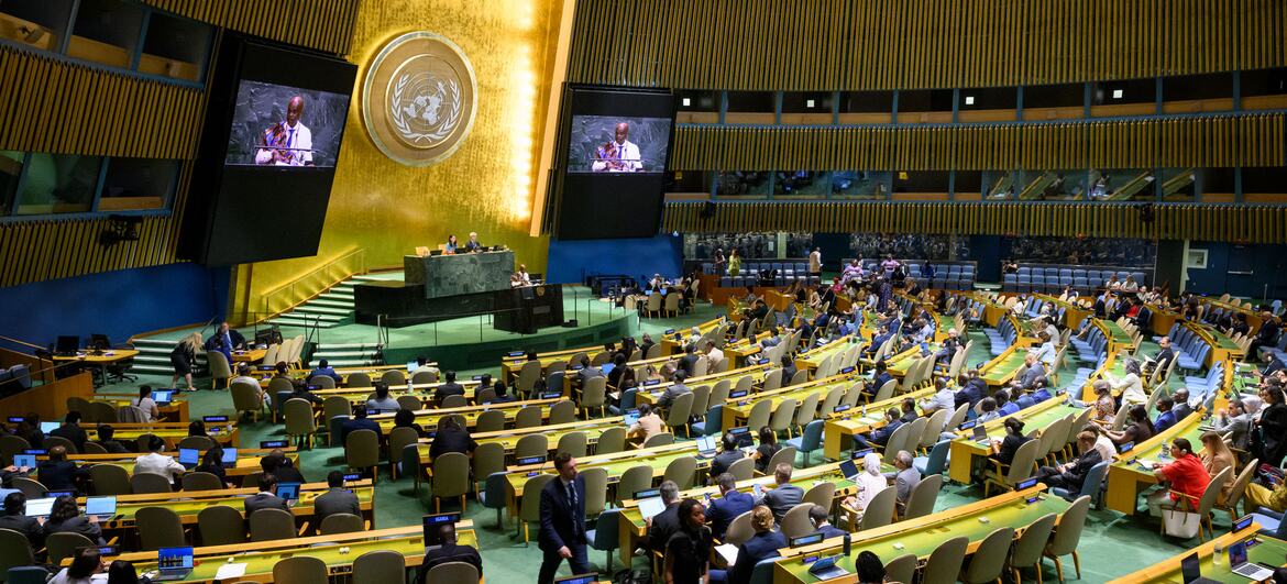 The United Nations General Assembly plenary hall in New York is filled with delegates seated at curved desks, facing a central podium beneath the UN emblem on a gold wall.