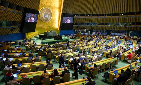 تمتلئ قاعة الجلسات العامة للجمعية العامة United Nations في نيويورك بالمندوبين الجالسين على مكاتب منحنية، يواجهون منصة مركزية أسفل شعار الأمم المتحدة على جدار ذهبي.