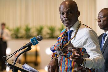 A Togolese man in traditional woven fabric and a white shirt speaks at a UN-branded microphone during an official meeting.