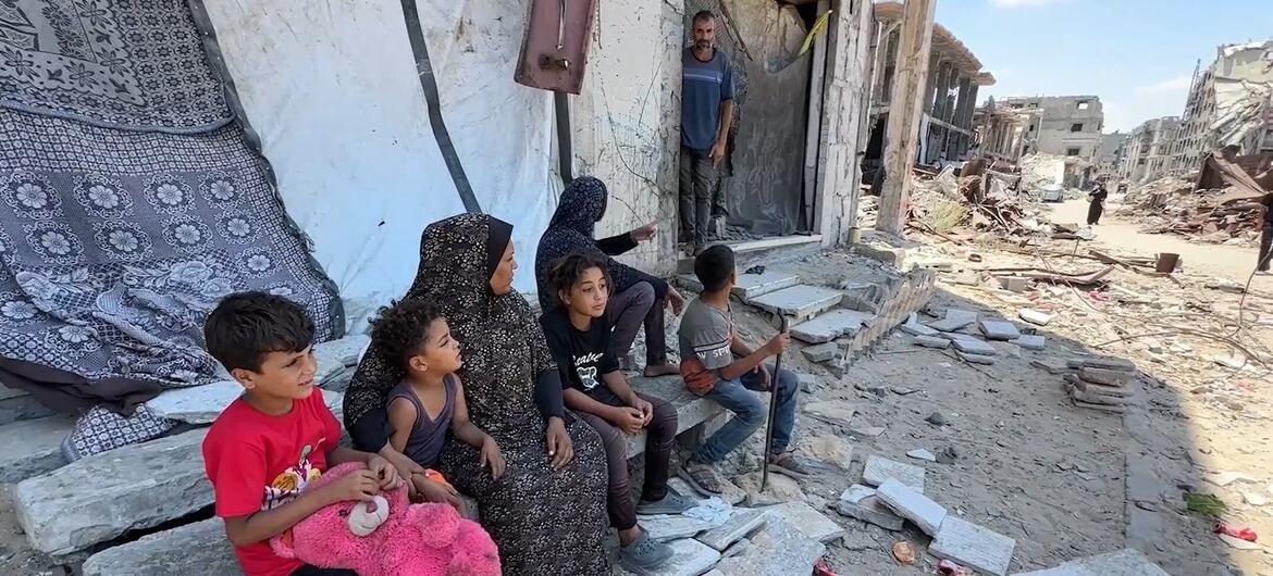 A Palestinian family, including a woman in a headscarf and four children holding a pink teddy bear, sits on concrete slabs amid rubble outside a damaged building in Gaza. A man stands in the doorway behind them as destroyed structures stretch into the background.