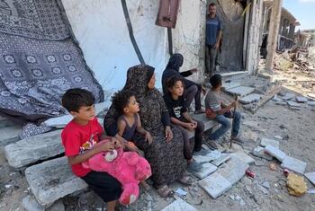 Una familia palestina, compuesta por una mujer con velo y cuatro niños que sostienen un osito de peluche rosa, se sienta sobre losas de hormigón entre los escombros de un edificio dañado en Gaza. Un hombre permanece de pie en la puerta detrás de ellos, mientras las estructuras destruidas se extienden hasta el fondo.