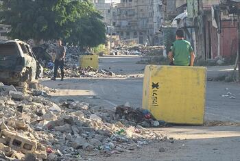 Un homme en chemise verte est assis sur un grand muret en béton jaune marqué du mot hébreu « jaune » et d'un symbole noir, dans une rue de Gaza. Un autre homme se tient à proximité. La scène est entourée de décombres, d'immeubles détruits, d'une voiture calcinée à gauche et d'une berline argentée intacte à droite.