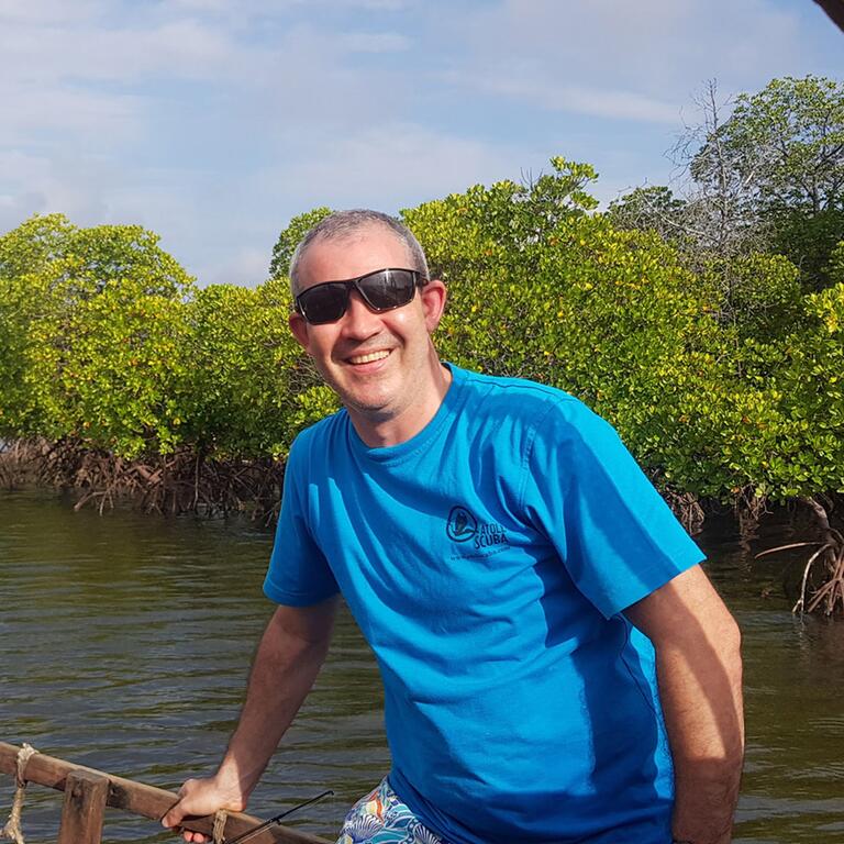 Gabriel Grimsditch, un hombre con camiseta azul y gafas de sol, está de pie en una barca de madera que navega por un canal tranquilo bordeado de densos manglares verdes.