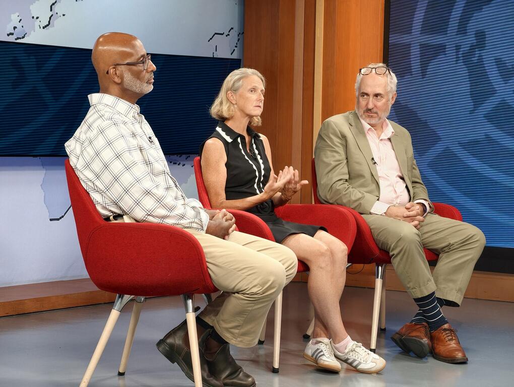 Three guests—two men and a woman—sit in red armchairs on a television studio stage during a panel discussion, with a woman gesturing as she speaks.