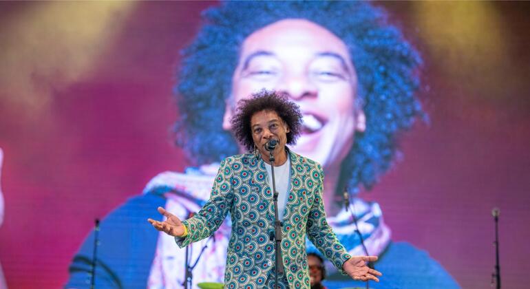 Stewart Sukuma se apresenta no palco em um evento da ONU News, vestindo um terno estampado em verde e azul, com os braços estendidos em frente a um microfone, enquanto um grande telão de LED atrás exibe uma imagem em close de seu rosto sorridente.