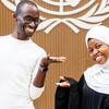Mwanamume Mweusi mwenye tabasamu akiwa amevaa sweta nyeupe na miwani na mwanamke Mweusi mwenye tabasamu akiwa amevaa hijab nyeupe na abaya nyeusi akiwa amesimama kando kando akifanya ishara za mikono wazi mbele ya ukuta wa mbao wenye nembo UNESCO .