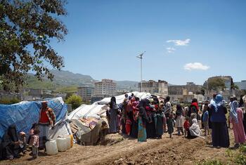 Group of displaced Yemeni civilians, mostly women and children in traditional dress, gather near blue tarp shelters and water containers on a dirt field with damaged buildings and mountains in the background.