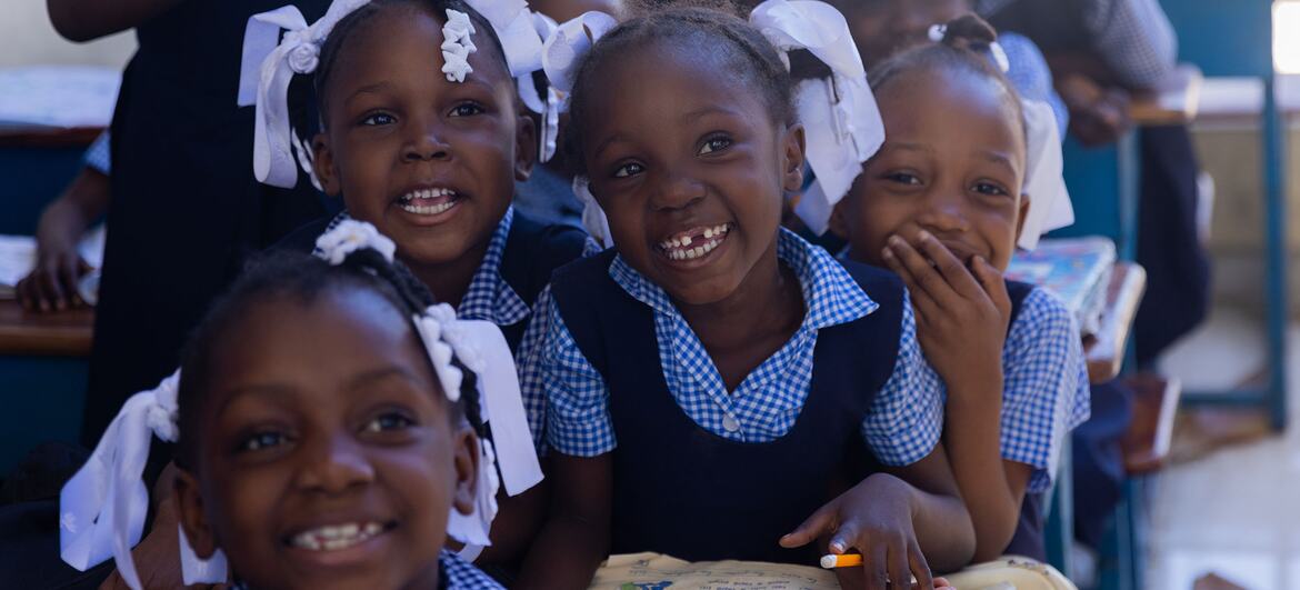 Young Haitian girls in blue and white checkered school uniforms with white hair bows smile at the camera inside a classroom in Delmas 45.