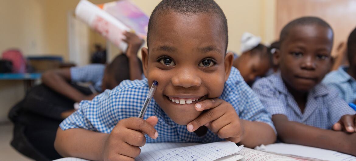 A smiling seven-year-old Haitian boy in a blue checkered shirt sits at a desk in a classroom holding a pen over an open notebook, with other students visible behind him.