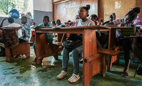 A group of young Haitian students sits at wooden desks in a classroom, taking notes and studying.